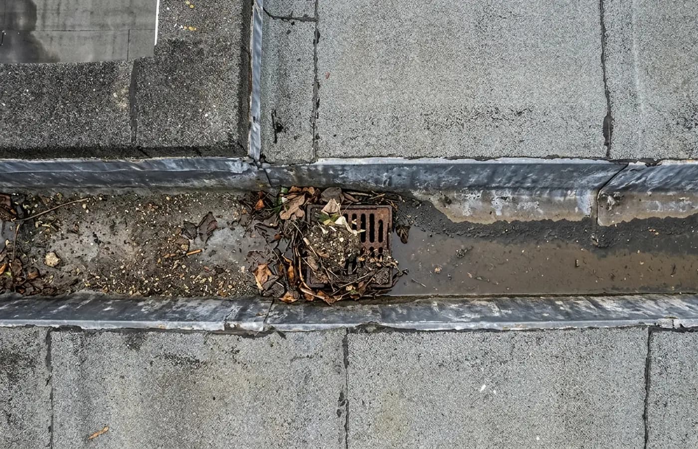 A parapet gutter on a commercial roof silted with grit and leaf litter, with a blocked outlet and a tide mark of standing water